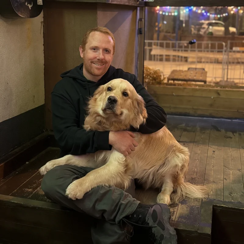 Curtis sitting on a bench with Whiskey the golden retriever, fairy lights and the street behind