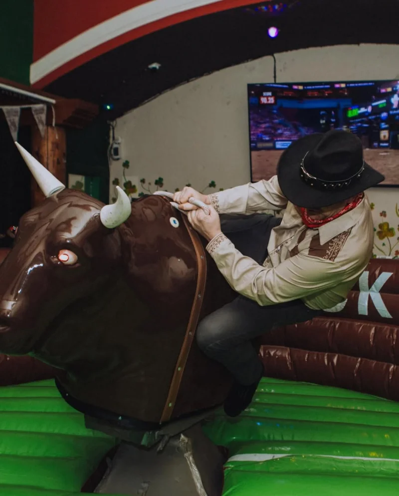 Cowboy on the mechanical bull at the Classic, green inflatable surround, TV behind showing pro bull riding